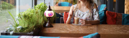 girl in a dress sitting at a table in a cafe and drinking coffee, waiting for a meeting, a date..dressed in a white dress with blue and pink flowers, sits by the window at the carved table, on which the lamp is burning ...の写真素材