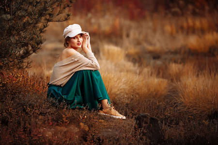 beautiful girl in a cap and coat with a bare shoulder on the background of autumn walk green skirt, sittingの写真素材