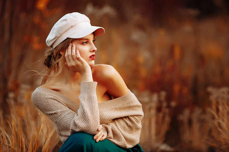 beautiful girl in a cap and coat with a bare shoulder on the background of autumn walk green skirt, sittingの写真素材