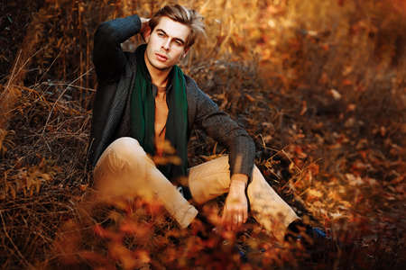 a man, a model in a gray jacket, a green scarf and beige pants and a sweater sits on the grass in autumn, among the autumn trees. posing. thoughtfulness and sadness. male powerの写真素材