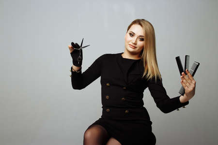 young woman with beautiful blonde hair look at scissors in one hand studio shot gray background sits on a chair holds three combsの写真素材