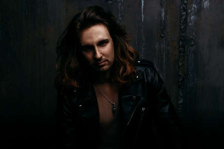 a brutal man in a black, leather jacket, with long hair, sits on a chair, against the backdrop of a...