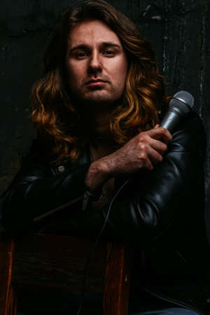 a brutal man in a black, leather jacket, with long hair, sits on a chair, in hands with a gray microphone. against the backdrop of a black wall. singer, radio, performance, portrait.の写真素材