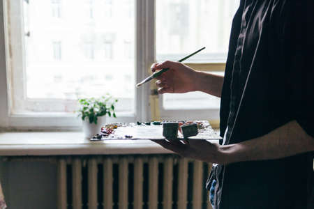 the artist holds a palette with paints and a brush and is going to paint on canvas. a man in jeans and a black shirt. hobby. across from the windowの写真素材