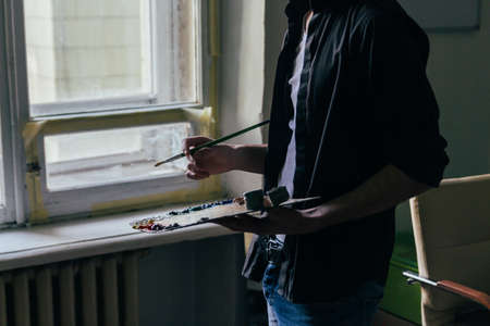 the artist holds a palette with paints and a brush and is going to paint on canvas. a man in jeans and a black shirt. hobby. across from the windowの写真素材