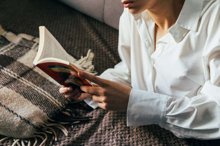 girl reading a book in a warm blanket. woman lies, lay down on the bed, a blanket, a sofa, a couch and holds a book. girl reading a book. education, recreation, training, self-improvement, scienceの写真素材