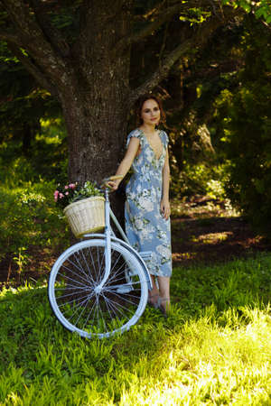 portrait of a beautiful girl in the forest, holding a bike with a basket of flowers, behind the rays of the sun, a blue flowered dress, summer walk relaxationの写真素材