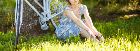 portrait of a beautiful girl in the forest, sitting on the grass, next to the bike, with a basket of flowers, behind the rays of the sun, a blue flowered dress, summer walk. relaxationの写真素材