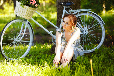 portrait of a beautiful girl in the forest, holding a bike with a basket of flowers, behind the rays of the sun, a blue flowered dress, summer walk relaxationの写真素材