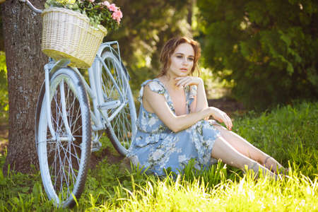 portrait of a beautiful girl in the forest, sitting on the grass, next to the bike, with a basket of flowers, behind the rays of the sun, a blue flowered dress, summer walk. relaxationの写真素材