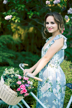 portrait of a beautiful girl in the forest, holding a bike with a basket of flowers, behind the rays of the sun, a blue flowered dress, summer walk relaxationの写真素材
