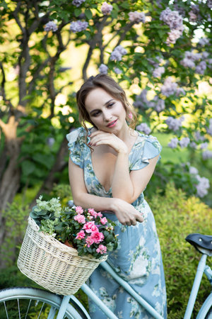 portrait of a beautiful girl in the forest, holding a bike with a basket of flowers, behind the rays of the sun, a blue flowered dress, summer walk relaxationの写真素材