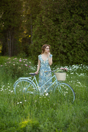 portrait of a beautiful girl in the forest, holding a bike with a basket of flowers, behind the rays of the sun, a blue flowered dress, summer walk relaxationの写真素材