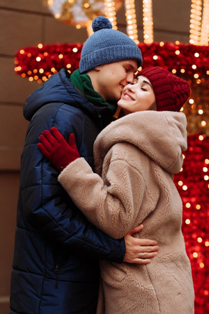 romance couple in love, girl and boyfriend. the girl with closed eyes kisses the guy in the cheek, the man smiles. burgundy hat and gloves, winter. on the background of lightsの写真素材