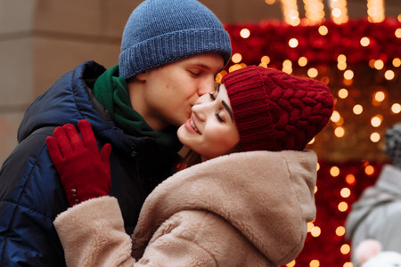 romance couple in love, girl and boyfriend. the girl with closed eyes kisses the guy in the cheek, the man smiles. burgundy hat and gloves, winter. on the background of lightsの写真素材