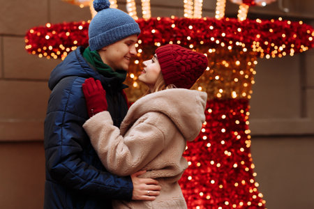 romance couple in love, girl and boyfriend. the girl with closed eyes kisses the guy in the cheek, the man smiles. burgundy hat and gloves, winter. on the background of lightsの写真素材