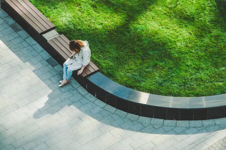 Frustrated Caucasian woman with the phone and cigarette sitting on the wooden bench in the garden. Birdeye copter top aerial viewの写真素材