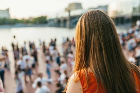 A girl with long hair in a red dress from the back looks at the crowd of dancing people on the middle plan. Selective focus macro shot with shallow DOFの写真素材