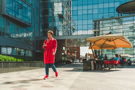 MOSCOW, RUSSIA - AUGUST 31, 2017. A happy girl in a red coat, blue jeans and red sneakers walks across the square between the office building and looks at the phone. In the background there are tables of a summer cafeのeditorial素材