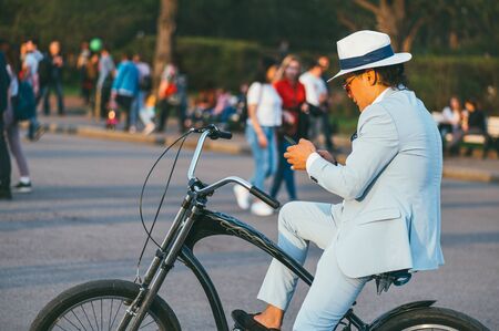 Moscow, Russia - may 1, 2018. Fashionable businessman in a hat, light jacket on a Bicycle, looking at the phone, against the background of a crowd of walking people In the Gorky Parkのeditorial素材