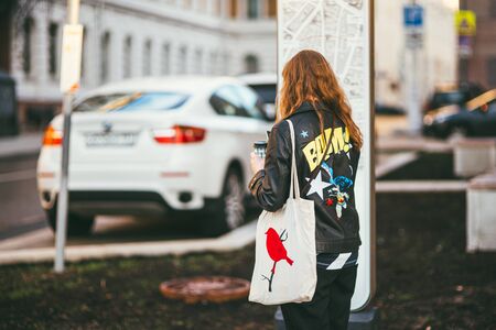 Moscow, Russia - may 25, 2017. A young woman in a black leather jacket with a bright yellow text print and a white bag with a red bird stands at an intersection near the road and calls a taxiのeditorial素材