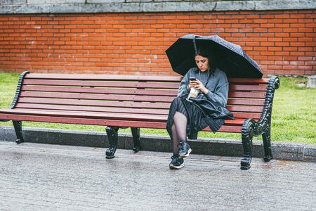 Moscow, Russia - JULY 7, 2017. A single woman in a black skirt and a gray jacket under a black umbrella sits on a bench and looks at the phone. Rainy day in Moscowのeditorial素材