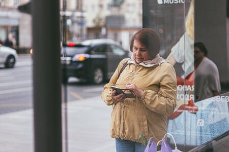 Moscow, Russia - JULY 7, 2017. A senior woman stands behind the glass of a bus stop and searches for something on her phone. Next to the bench are bags with purchases. And in the background you can see the road with carsのeditorial素材