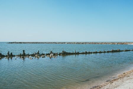 An old ruined pier with wooden logs sticking out of the water from the sea surface. Sandy beach in the foregroundの写真素材