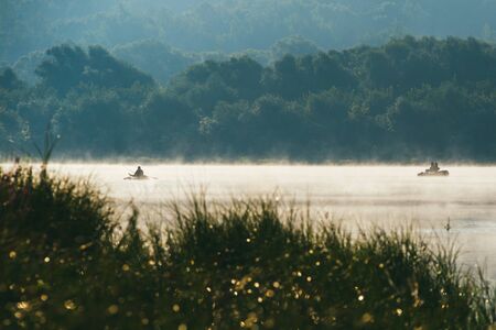 Fishermans fishing on a boat. Early morning spring landscape with river in the foreground and a green bank with bushes and trees in the background. The distant plan is drowned in a thick morning fogの写真素材