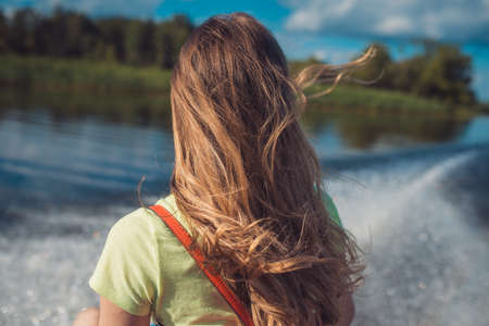 a young woman with long hair rides a fast boat on the water surface of the lake. Back view. Against the background of a white foam trail from the propeller and green shores receding into the distanceの写真素材