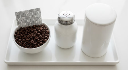 A trio of breakfast essentials, showcasing dark roasted coffee beans in a bowl, a classic salt shaker, and a white cup, artfully arranged on a clean tray.の素材