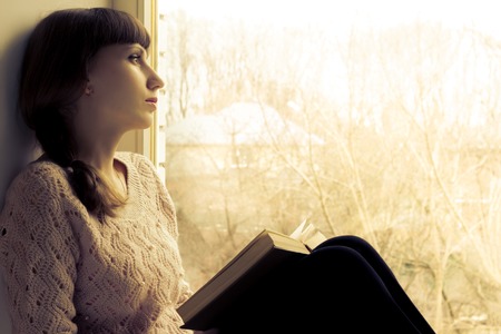 Young woman reading book near the window. Warm toned imageの写真素材