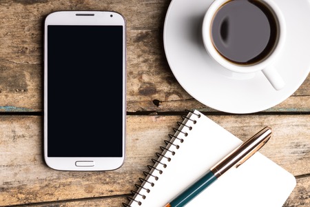 Smartphone with notebook and cup of strong coffee on wooden background. Cell phone with writing set with espressoの写真素材