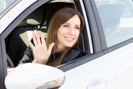 Young smiling woman greeting with hand from car. Cheerful caucasian girl welcome somebody sitting in...