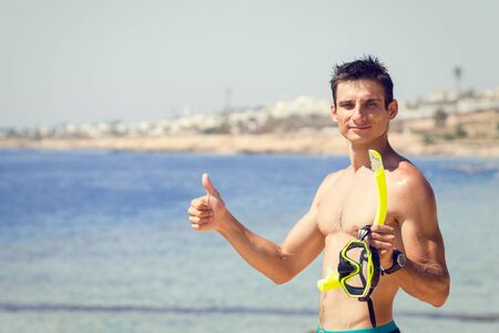 Young man with mask and snorkel shows thumbs up at the sea beach. Summer vacation and snorkeling background with copy spaceの写真素材
