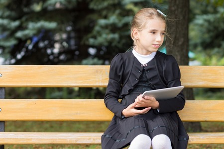 Cute young student girl with tablet sitting in the park. School child resting on the bench outdoors. Copy space area in the leftの写真素材