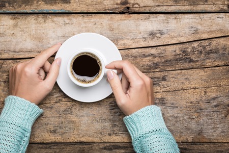 Woman's hands in sweater hold a cup of strong coffee on wooden table. Coffee fan top view background...
