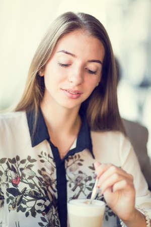 Young beautiful woman having her morning latte in restaurant. Gorgeous student girl stirring breakfast coffee in cafeの写真素材