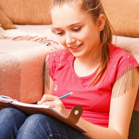 Young happy smiling teenager girl writing in diary notebook sitting on floorの写真素材