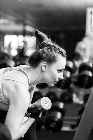 Young confident woman doing biceps curl exercise with dumbbells in fitness center. Slim girl training in the gym with dumbbells. Black and white imageの写真素材