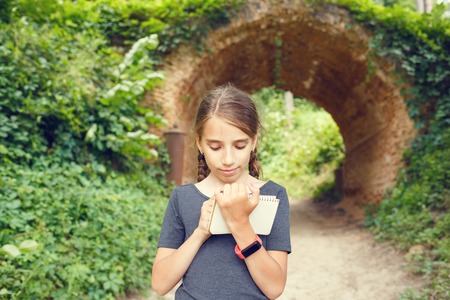 Small teenage girl with smartwatch drawing in notebook near beautiful old arch bridge in park. Cute hispanic travelling girl writing in diary on summer holidaysの写真素材