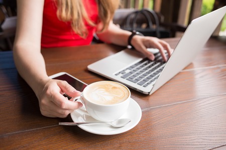 Young woman in red dress working with laptop in cafe holding cup of cappuccinoの写真素材