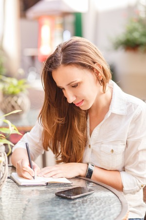 Young woman writing down into her notebook sitting at the table in restaurantの写真素材
