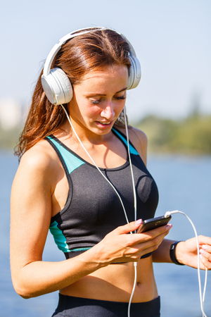Young sporty woman using smartphone on training . Smiling girl with headphones listening to music outdoor workout at summer morningの写真素材