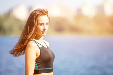 Portrait of young sporty woman resting after jogging in park near lake. Portrait of athletic girl in black top after fitness workout. Healthy lifestyle image with copyspaceの写真素材