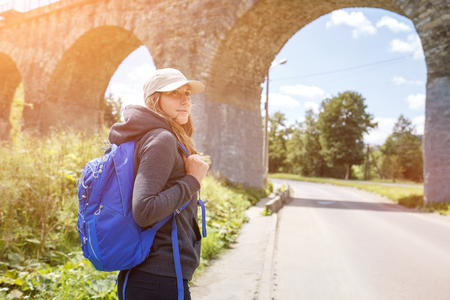 Young hitchhiker woman with backpack standing near old viaduct. Tourist backpacker girl in scenery country sideの写真素材