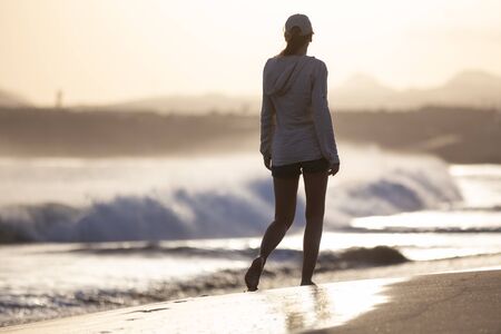 Young woman on vacation walking along ocean beach with great waves. Slim girl walking at seacoast in the eveningの写真素材
