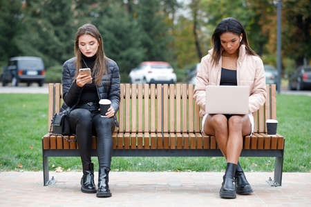 Two different busy women sitting on the bench in the parkの写真素材