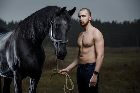 Young bearded man with naked torso standing with horse at the countrysideの写真素材
