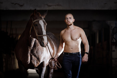 Young strong bearded man with naked torso walking with horse in the stableの写真素材